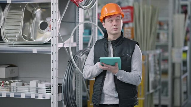 Concentrated male supervisor in hart hat standing between rows in hardware store examining goods on shelves. Portrait of focused confident Caucasian man working in repair supplies shop.