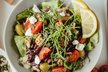 Healthy fresh salad, cucumber, tomato, spinach, arugula and olives and cheese in a plate on a white table, pumpkin, walnut and sunflower seeds with yellow lemon. Food background. From above close up