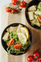 Two black bowl of rigatoni pasta with arugula, green peas, basil and cherry tomato served on wooden board. Italian food concept, from above close up