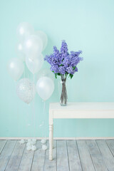 A bouquet of lilacs in a high vase on the table against the background of a blue wall. White balloons for the holiday.