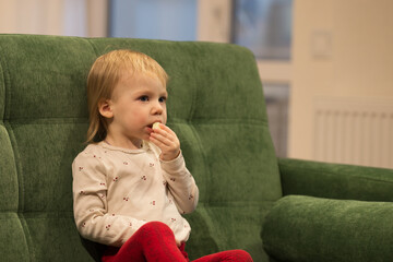 the child is sitting on the sofa in his house, holding an apple in his hand and putting it in his mouth to eat