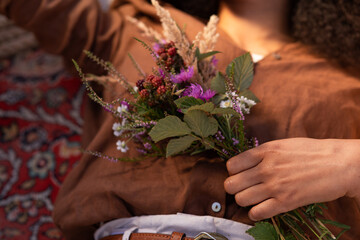 Eine Frau liegt auf dem roten Teppich, sie trägt eine braune Bluse, auf ihrer Brust in der Hand liegt ein Blumenstrauß vom Feld. Die Sonne scheint auf das Gesicht