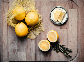 Lemons spilling out of a yellow net bag along with halved and sliced lemons, on a wooden plank