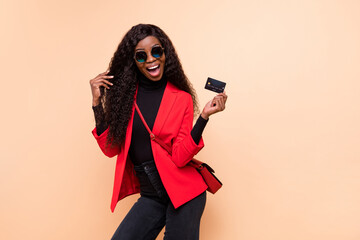 Photo of amazed happy dark skin woman hold credit card wear bag sunglass isolated on beige color...