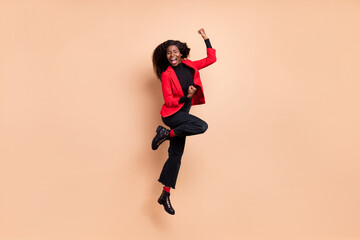 Fototapeta na wymiar Photo of excited lucky curly dark skin woman dressed red blazer jumping rising fists isolated beige color background