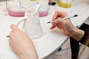 A woman applies a pencil sketch on a white ceramic jug before applying glazed paints. The concept of manual creativity, leisure, hobby