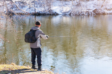 A fisherman with a fishing rod and a backpack catches fish on the bank of a snow-covered river in...