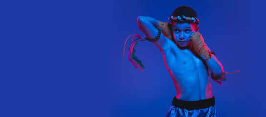 Young boy thai boxer posing over blue background in neon light.