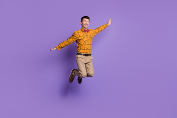 Fototapeta na wymiar Full length photo of funky carefree young man wear plaid shirt bow tie jumping arms sides isolated violet color background