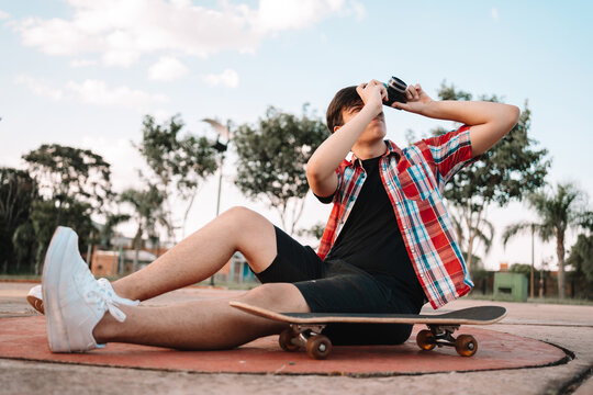 Young teenage man sitting on a skateboard outdoors taking pictures with a camera.