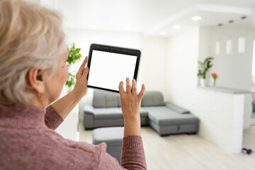 Senior woman at home standing at kitchen holding digital tablet controlling smart home system back view checking cameras close-up