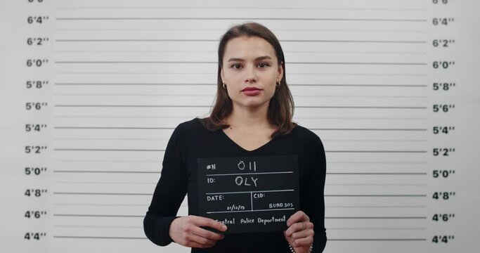 Mugshot of young attractive woman with holding sign for photo in police department. Crop view of pretty brunette female criminal posing, raising head and looking to camera.