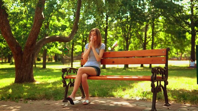 young woman in denim shorts and casual t-shirt has seasonal allergy sitting on the bench in summer park