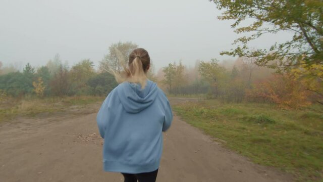 Slow motion tracking rear view of young woman in hoodie jogging along path in park on foggy autumn day