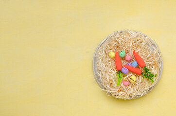 Top view of plate and easter nest on it, decorative eggs and carrots as a symbol of happy easter on the yellow surface.Empty space