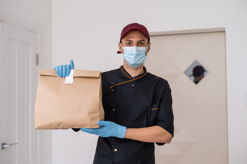 Fototapeta na wymiar Young delivery man in medical gloves and protective face mask holding paper bags in hands. Man courier with shopping bag. Safe online delivery from supermarket to home. COVID-19 coronavirus protection
