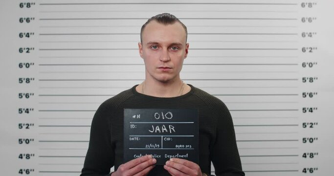Portrait of male criminal person holding sign for photo in police department. Crop view of serious man posing, raising head and looking to camera. Concept of mugshot and crime.