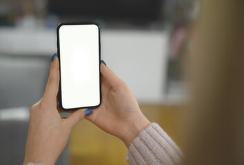 Back view close up of a woman hand holding and using smart phone with white blank empty screen sitting on a bed at home