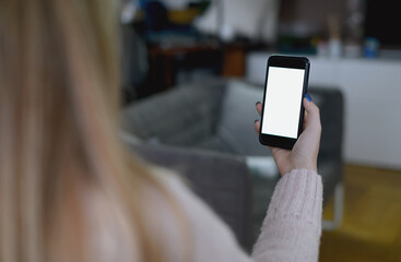 Back view close up of a woman hand holding and using smart phone with white blank empty screen sitting on a bed at home