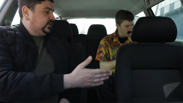 Two men with beards and mustaches are sitting in car in passenger seats, one on side and other behind driver, talking to each other and waiting for taxi driver to come and take them to destination.