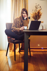 A pregnant woman sitting at the dining table, drinking coffee and looking at the laptop.