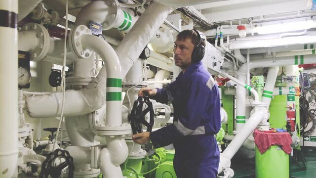 concentrated motor mechanic rotates black valve standing in engine room of freight vessel among various pipelines