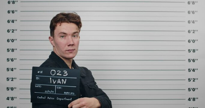 Side profile mugshot of millennial man with red hair turning head and looking to camera. Arrested guy holding sign for photo while standing in front of police metric lineup wall.