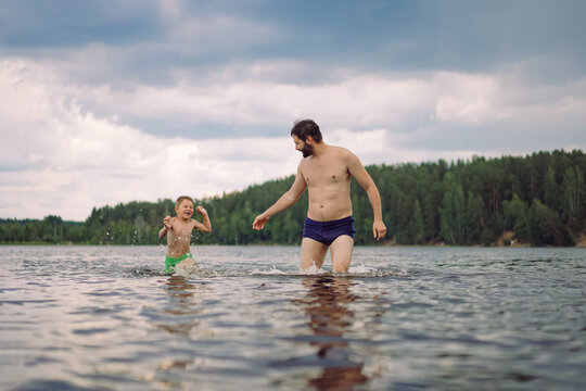 Young Bearded Caucasian Man And His Little Cute Son Running In The Lake In Forest And Splashing. Boy Is Chasing His Father Image With Selective Focus