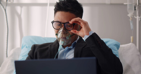 Tired sick businessman lying in hospital bed wearing suit and oxygen mask and working on laptop