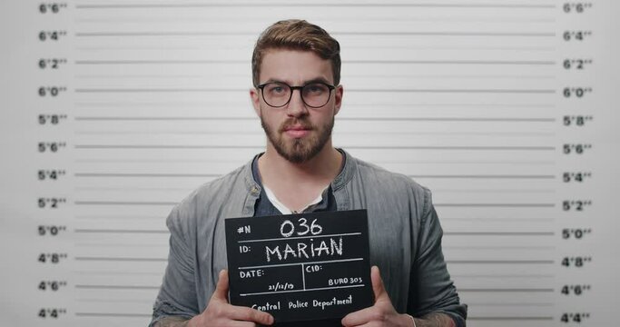 Portrait of tough guy with beard and mustache holding sign while being photographed in police station. Male person with in glasses posing and looking to camera. Concept of mugshot.
