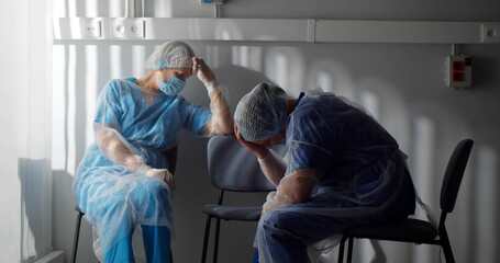 Tired surgeons in sterile gown and safety mask sitting on chairs in hospital room and resting