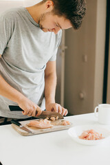 Young man cutting chicken fillet with knife on wooden board in modern white kitchen. Home cooking