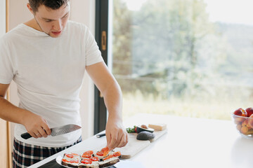 Young man putting tomato slices on bread and making avocado toasts in modern white kitchen. Healthy