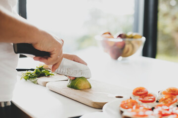 Man cutting avocado and making toasts with tomato, arugula, cheese in modern kitchen. Home cooking