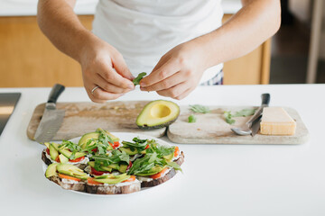Young man making toasts with avocado, tomato, arugula, cheese in modern kitchen. Home cooking