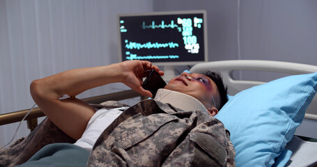 Injured asian soldier with neck collar lying in hospital bed and talking on smartphone