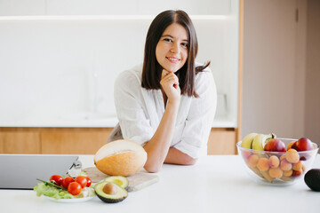 Young woman sitting at white countertop with fresh fruits and vegetables in modern white kitchen