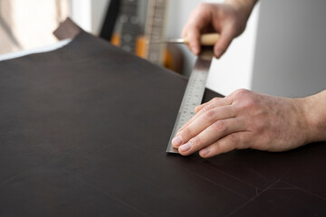 Men s hand holding a awl and metal ruler and working on a leather wallet in his workshop. Working process with a brown natural leather. Craftsman holding a crafting tools and put marks on a leather.