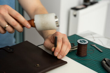Mens hand holding a leather carving hammer and steel chisel hole punch and makes a leather wallet in his workshop. Working process with a brown natural leather. Craftsman holding a crafting tools.