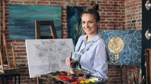Portrait of female artist mixing color paints with art brush on the palette. Young beautiful painter at the studio preparing for painting a still life picture with sunflowers.