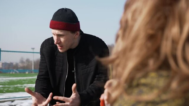 A young gopnik man in a black jacket and rolled-up hat speaks emotionally to a woman sitting opposite and gesturing with his hands on playground. A people with long hair gives him painful five on arm