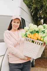 Woman with a delivery truck carries a large basket of flowers. Deliver flowers. Supply garden plants. Gardener company brings flowers home. Delivery woman carries flowers