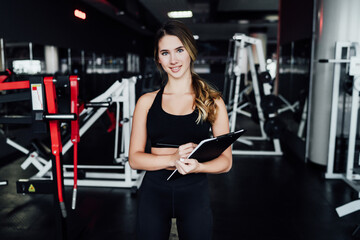 Fototapeta na wymiar Fitness blonde girl personal fitness trainer with workout plan in hand, smiling and looking at camera, she is in the middle of modern gym