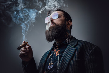 Elegant man in retro style poses in dark background smoking cigar. Portrait of a bearded guy with...