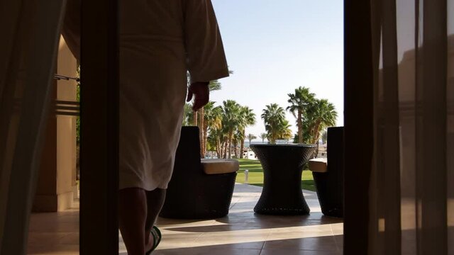a man in a white robe and with a tablet leaves the room on the terrace overlooking the sea, sits in an armchair and scrolls in the tablet, curtains in the foreground, palm trees in the background