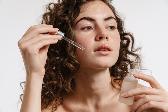 Half-naked curly woman applying face serum while looking at camera
