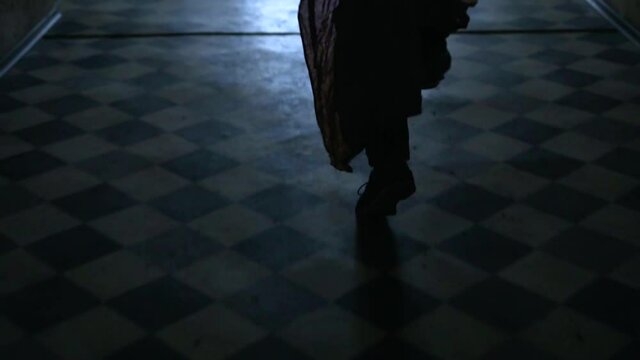A silhouette of a man running across the tile floor along a dark corridor to the light at the end and hurrying. A man in baggy Indian pants. Rear view. Close-up of legs