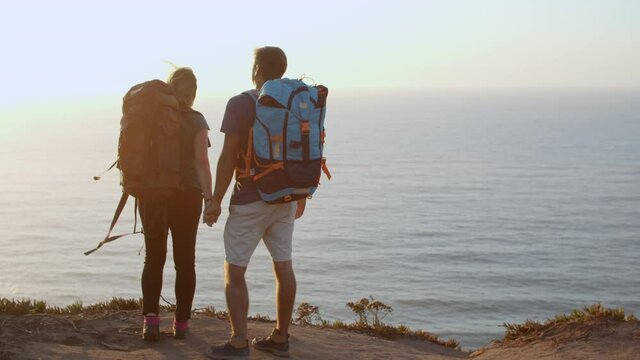 Happy couple with camping backpacks walking on mountain path along cliff, enjoying view of sea and sunset. Dolly shot. Travel activity concept