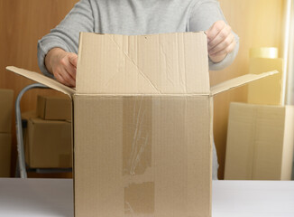 Fototapeta premium woman in a gray sweater is packing brown cardboard boxes on a white table, behind a stack of boxes