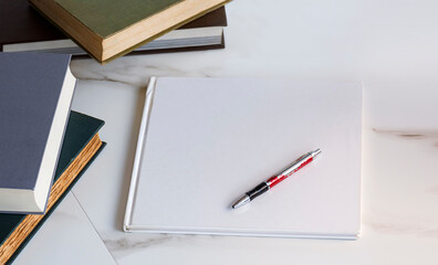 Pen on blank white book cover with various old textbooks stack on white marble desk 